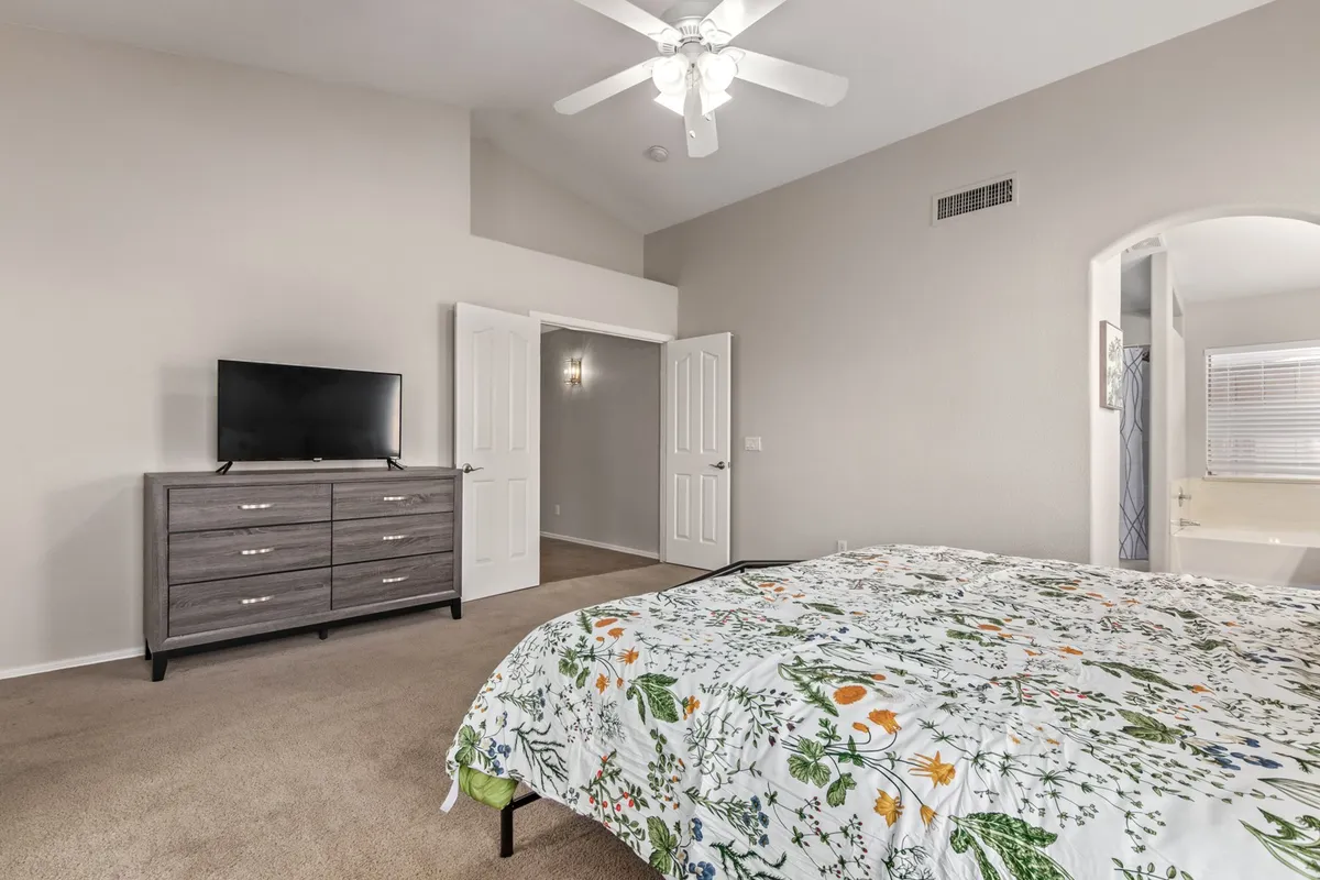 A cozy bedroom with floral bedding, a dresser with a TV, and open doors leading to another room. The decor is soft and inviting, with a focus on relaxation and comfort, enhanced by the subtle presence of the TV—perfect for unwinding.