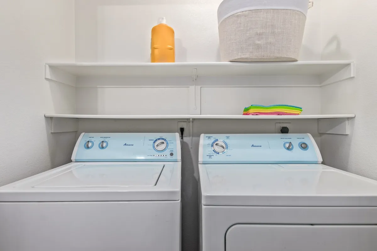 A functional laundry room featuring a modern washing machine and dryer, organized shelves stocked with cleaning supplies, and a clean, uncluttered workspace. The space is bright and practical, designed for efficiency and ease of use.