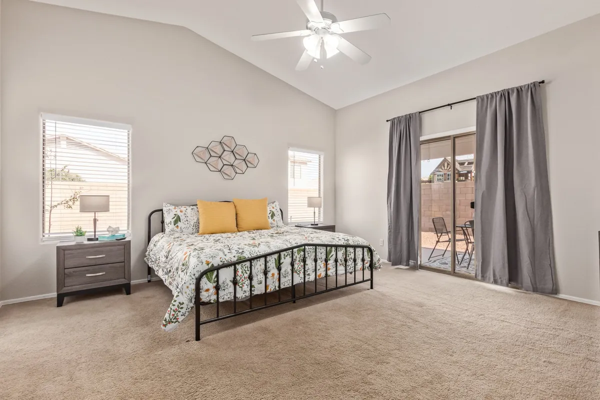 A serene bedroom scene featuring a bed with floral bedding, yellow accent pillows, a nightstand with a lamp, large windows, and a sliding glass door that lets in abundant natural light. The calm, inviting mood makes it ideal for relaxation and unwinding.
